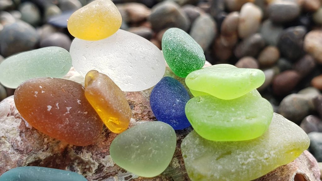 unusual coloured seaglass, in yellow, amber, lime green, sage, cobalt, kelly green on top of a pacific abalone shell on a beach with tiny pebbles
